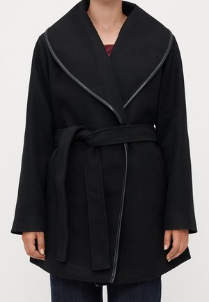 Czarny płaszcz z paskiem i szerokimi klapami obszytymi skórzaną lamówką, noszony na ciemnych dżinsach, pokazujący linię szyi i tułów stojącej osoby.