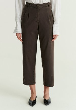 Vrouw die donkerbruine cropped broek met plooien draagt, gecombineerd met een wit hemd met lange mouwen en zwarte puntige schoenen.