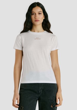 Jonge vrouw met lang donker haar, gekleed in een wit T-shirt met de tekst "W.11 NOTTING HILL" en zwarte jeans met bloemmotief uitsnijdingen.