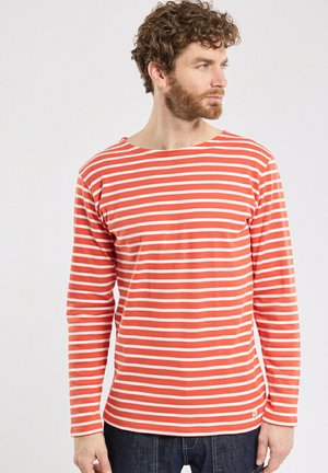 Homme aux cheveux bouclés et à la barbe, portant un t-shirt à manches longues rayé horizontalement rouge et blanc, regardant vers sa droite.