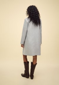 Manteau en laine gris à coupe droite, manches longues, ourlet légèrement fuselé, associé à des bottes de cow-boy marron avec des détails embossés.