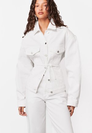 Kobieta o długich kręconych włosach, ubrana w białą, oversize'ową kurtkę dżinsową, zaciśniętą w talii, oraz pasujące białe spodnie, na jednolitym tle.