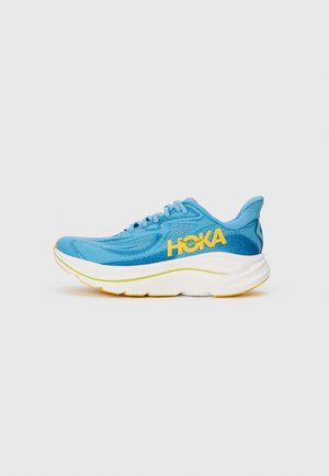Blue athletic shoe with mesh upper, yellow accents, thick white outsole, and textured design, featuring HOKA logo on the side.