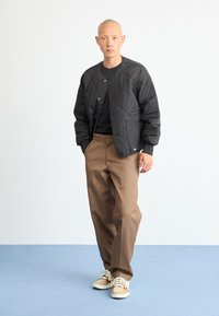 Bărbat cu cap ras, îmbrăcat într-o jachetă neagră matlasată, cămașă neagră, pantaloni maro și adidași bej, stând împotriva unui fundal alb și albastru simplu.