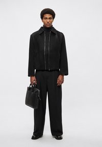 Crna skraćena jakna s prednjim patentnim zatvaračem i okovratnikom, uparena s crnim hlačama širokih nogavica. U jednoj ruci drži tanku crnu kožnu torbicu.