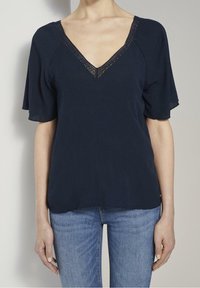 Donna che indossa una blusa blu navy con scollo a V e rifiniture in pizzo, con maniche corte, abbinata a jeans azzurri, in piedi contro uno sfondo grigio.