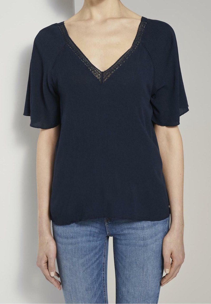 Donna che indossa una blusa blu navy con scollo a V e rifiniture in pizzo, con maniche corte, abbinata a jeans azzurri, in piedi contro uno sfondo grigio.