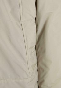 Beige isolerad jacka i tyg med slät yta. Har synliga sömmar och en sömlös ficka på vänster sida.