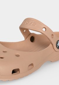 Sabot style Crocs marron clair avec des trous de ventilation et une bride ajustable assurant le maintien du talon, présenté sur un fond uni.