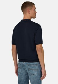 Jeune homme aux cheveux courts portant une chemise noire à manches courtes et un jean bleu clair, debout de dos sur un fond uni.