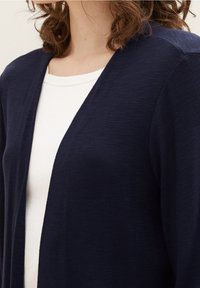 Granatowy kardigan o miękkiej fakturze, z przodem na otwarcie i dekoltem w kształcie litery V, noszony na białej dopasowanej bluzce.
