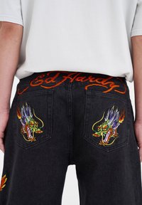 Černé džíny z denimu s barevným výšivkem draka na obou zadních kapsách a nápisem "Ed Hardy" vyšitým oranžově na pásku.
