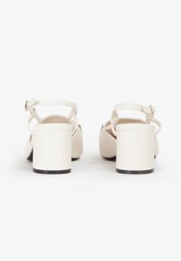 Sandalias de tacón blanco de cuero con tacón cuadrado, correa al tobillo y diseño cruzado, con una textura suave y suela negra.
