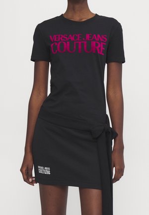 Osoba nosząca czarny top z krótkim rękawem z "Versace Jeans Couture" w kolorze różowym oraz dopasowaną czarną spódnicę z wiązanym detalem i białym logo.