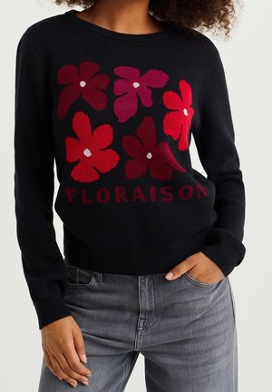 Pull noir avec six grandes fleurs rouges et bordeaux et le texte « FLORAISON », porté avec un jean gris, une main dans la poche.