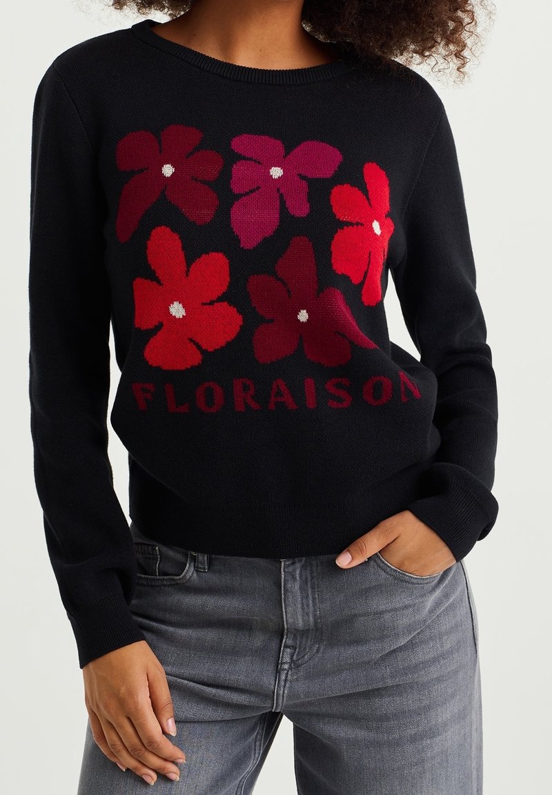 Pull noir avec six grandes fleurs rouges et bordeaux et le texte « FLORAISON », porté avec un jean gris, une main dans la poche.
