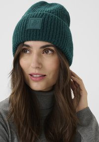 Teal rebrast knitev klobuk z teksturirano površino in kvadratno gumo oznako. Uporabljen z sivo turtleneck majico v nevtralnem ozadju.