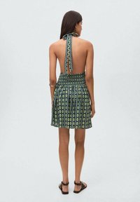 Vista posteriore di un vestito senza maniche con collo halter di colore verde scuro, con un motivo floreale giallo e bianco, dotato di vita arricciata e schiena scoperta.