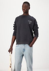 Mörkgrå crewneck sweatshirt med ribbstickade ärmar, prydd med en vit "GUESS"-logotyp på bröstet, tillsammans med ljusblå baggy jeans.