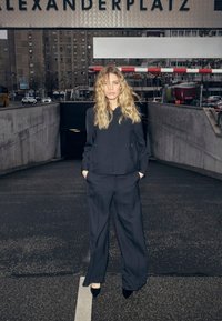 Costum negru în două piese cu jachetă cu fermoar și pantaloni largi. Materialul pare neted, purtat cu pantofi negri cu vârf ascuțit. Fundalul prezintă un cadru urban.