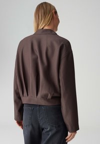 Braune, kurze Jacke mit glatter Textur, übergroßen Ärmeln und Schalkragen. Trägt man über dunklen Jeans aus Denim, was eine entspannte Passform zeigt.