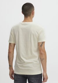 Tricou scurt cu mânecă scurtă de culoare crem deschis, cu o textură moale și decolteu rotund, având un design simplu. Vederea din spate arată tatuaje pe brațe.