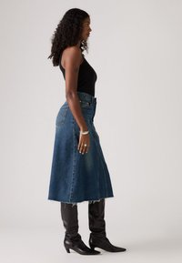 Denim midi suknja s rasparanim rubom, plave boje, visoki struk, uparena s crnim kožnim čizmama do koljena. Glatka tekstura kroz cijelu suknju.