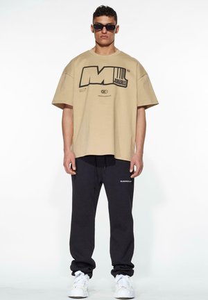 Beige oversized t-shirt with black graphic print, paired with dark jogger pants. The outfit features relaxed fit and white sneakers.