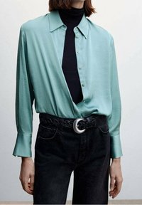 Lys turkise skjorte med krave, parret med en sort turtleneck og høj-taljede sorte jeans. Har et flettet sort bælte.