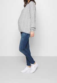 Harmaa palmikkoneulepusero, väljä istuvuus, yhdistetty sinisiin kapeisiin farkkuihin ja valkoisiin tennareihin. Yksinkertainen muotoilu, teksturoitu neule kohotetuilla kuvioilla.