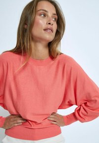 Koralröd tröja med rund halsringning och långa, batwingärmar. Mjuk, strukturerad tyg med ribbade muddar och nederkant. Positionerad mot en ljus bakgrund.