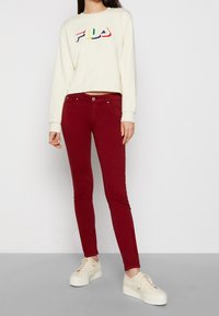 Bílá zkrácená mikina s vícebarevným logem FILA, spárována s těsnými vínovými kalhotami a béžovými platformovými teniskami.