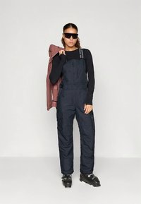 Žena v čiernych lyžiarskych overalových nohaviciach, dlhom rukáve, lyžiarskych topánkach, slnečných okuliaroch, drží fialovú bundu cez rameno na neutrálnom pozadí.