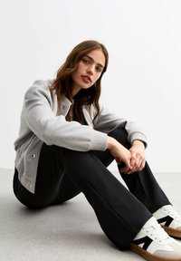 Grijze gebreide bomberjack met geribbelde manchetten, zwarte broek met een relaxte pasvorm en witte sneakers met zwarte accenten. Neutrale achtergrond.