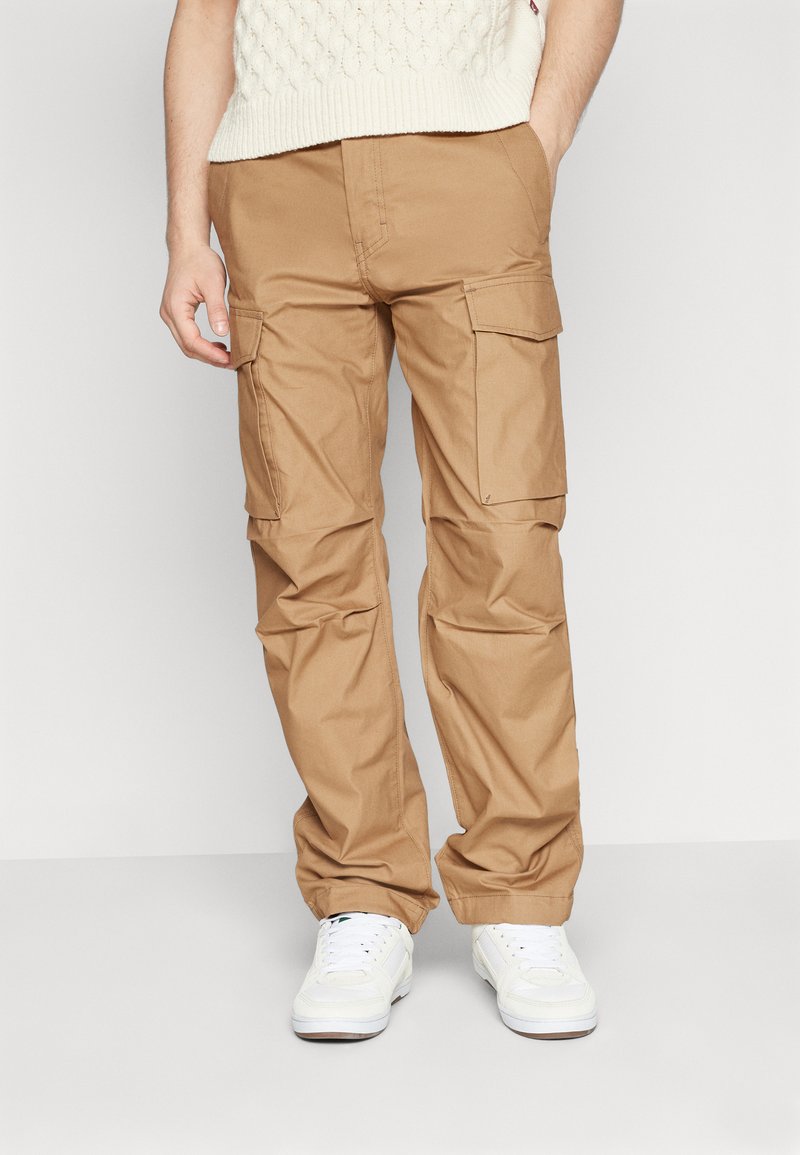 Beige cargo-bukser med flere store lommer, laget av bomullstoff. Har en avslappet passform og rette ben, kombinert med hvite sneakers.