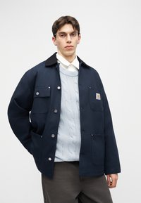 Mladý muž se brýlemi, v tmavě modré bundě přes světle modrý svetr s copánkovým vzorem a bílou košilí, stojící před jednobarevným pozadím.