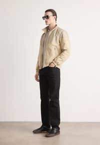 Casaco bege com parte superior texturizada, bolsos com fecho éclair e punhos elásticos, combinado com calças pretas e ténis escuros. Óculos de sol utilizados.