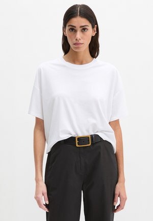 Vrouw met stijl donker haar die een effen wit oversized t-shirt draagt, gestopt in een high-waisted zwarte broek met een zwarte riem en gouden gesp.