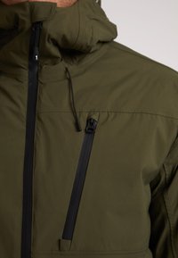 Grøn letvægtsjakke med høj krave, udstyret med en sort vandtæt lynlås og en lynlåslomme, glat tekstur og minimalistisk design.