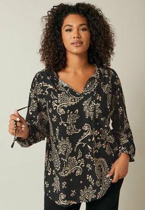 Zwarte blouse met een beige paisleypatroon, V-hals, lange mouwen en textuurstof. Het model houdt een strikdetail bij de kraag vast.