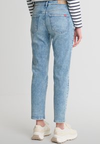 Gaiši zili džinsi ar augstu jostasvietu, taisnu kāju dizainu un divām aizmugures kabatām. Apvienoti ar bēšiem sporta apaviem. Daļēji redzama svītraina blūze.