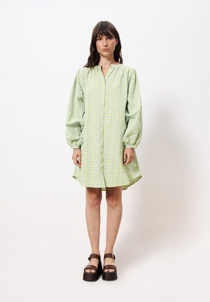 Robe à carreaux verts et blancs avec de longues manches bouffantes, fermeture à boutons sur le devant et coupe ample, associée à des chaussures à plateforme noires chunky.