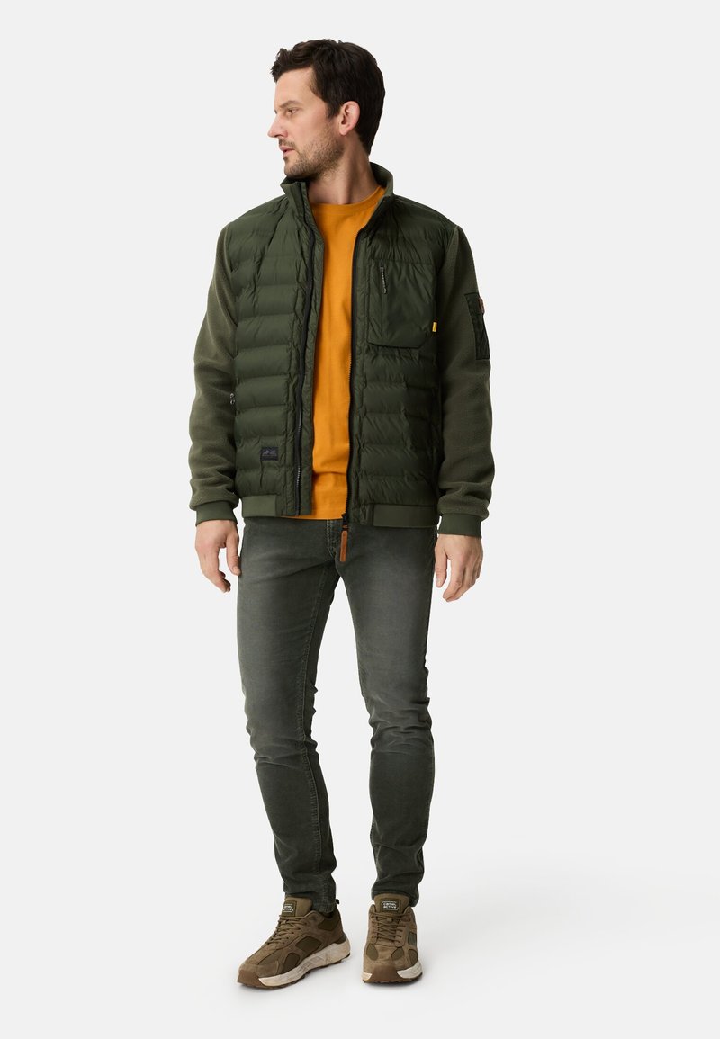 Mørkegrøn puff jakke med lynlås foran, kombineret med en orange skjorte og grå jeans. Brune sneakers fuldender outfittet.