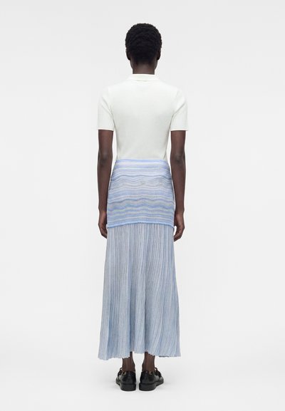 Άτομο που φοράει λευκό κοντομάνικο τοπ και μπλε πλισέ μάξι φούστα με πλεκτή μπλε ζώνη στη μέση, στέκεται μπροστά σε απλό φόντο.