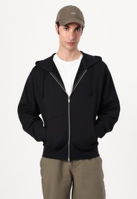 Un sweat à capuche zippé noir avec une capuche, doté d'une fermeture éclair argentée, porté sur une chemise blanche, associé à une casquette vert olive et un pantalon beige.