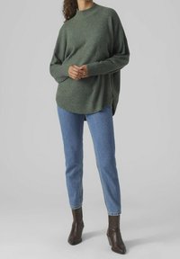 Femme portant un pull vert oversize, un jean bleu et des bottines marron foncé, debout devant un fond gris uni.