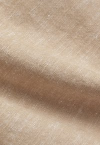 Tissu beige tissé avec une texture visible et de petites taches blanches, présenté en plis doux pour mettre en valeur les détails du matériau.