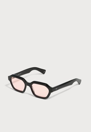 Schwarze geometrische Sonnenbrille mit hellrosa getönten Gläsern, die auf einer grauen Oberfläche liegt.