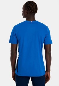 Camiseta azul de manga corta hecha de tejido suave. Presenta un cuello redondo con un pequeño acento rojo y blanco en la parte posterior. Diseño simple, sin patrones.