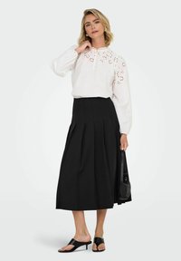 Blusa bianca con accenti ricamati, scollatura alta e maniche lunghe abbinata a una gonna plissettata nera e sandali neri con tacco.